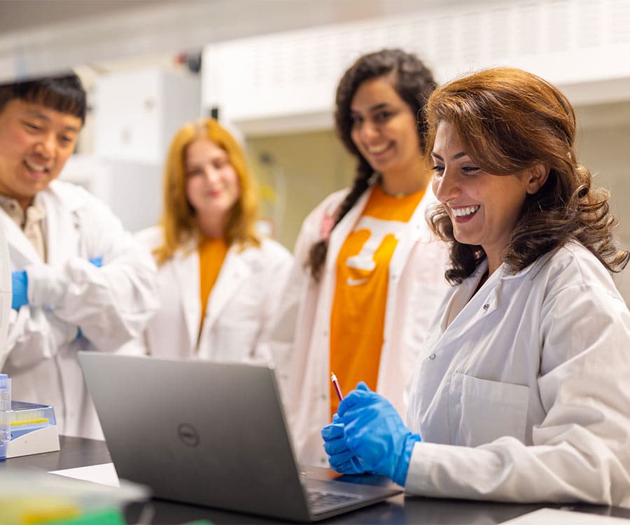 A UT researcher and a team of graduate students in white lab coats and blue gloves gathered around a laptop computer