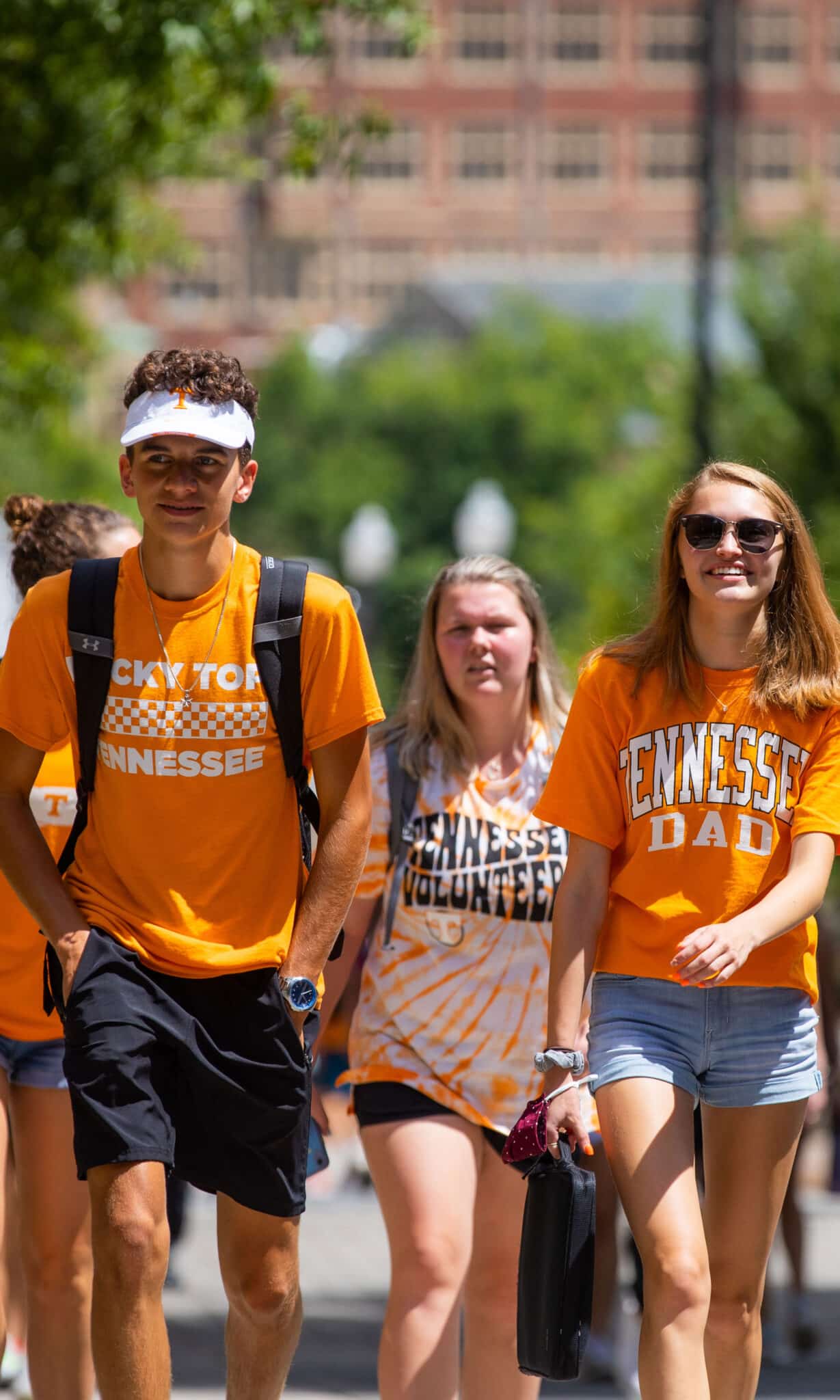 Three students walk across campus wearing orange UT t-shirts, hats, and sunglasses
