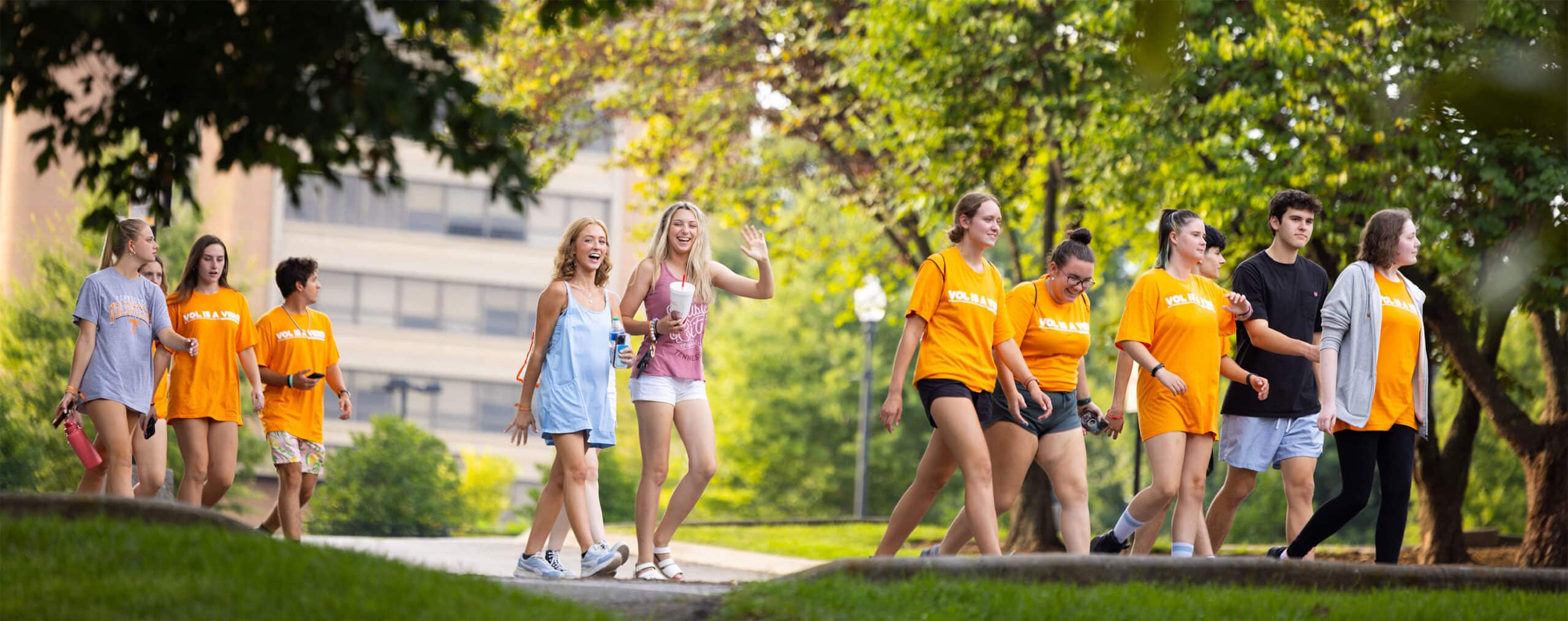 Students dressed in orange Vol is a Verb t-shirts walk across campus - one is waving at the camera