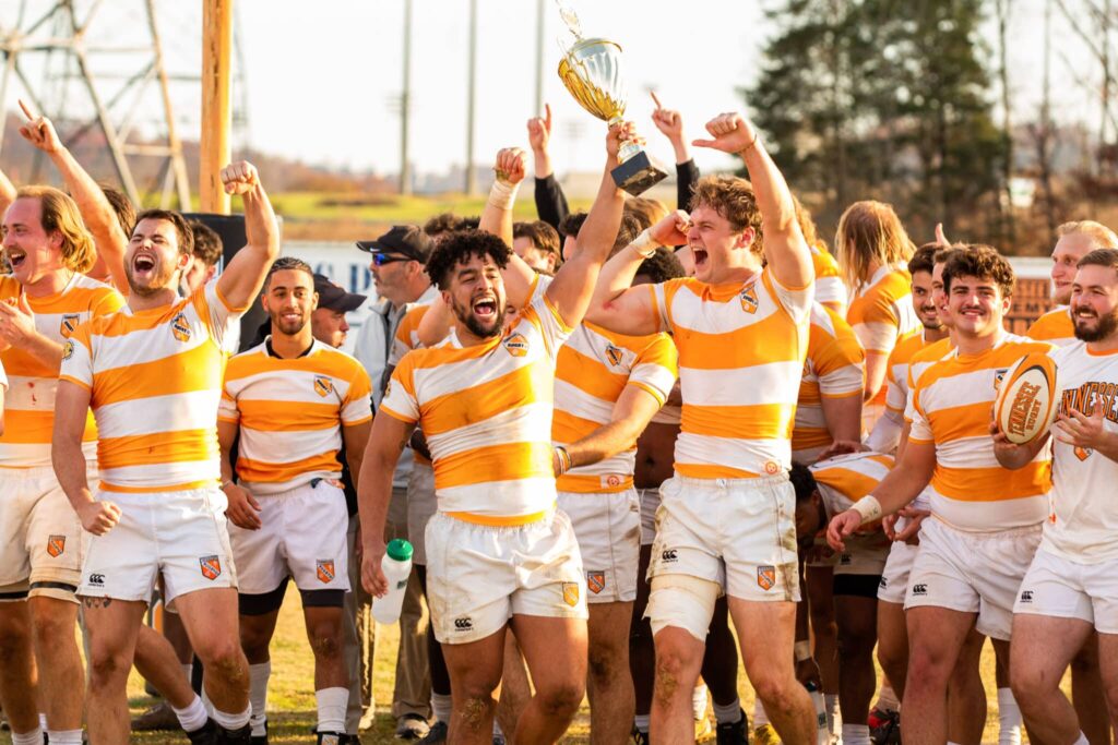 The men's intercollegiate rugby team cheers, wearing orange and white striped rugby shirts and white shorts