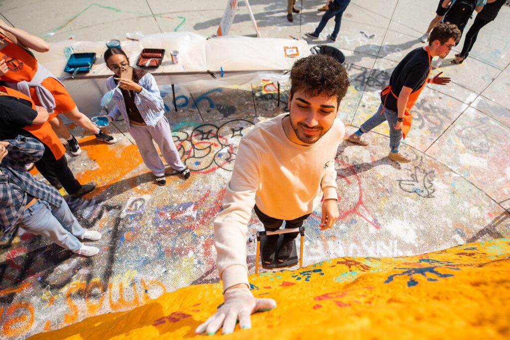 A student wearing a white shirt and glove places his handprint on the Rock, which is painted orange