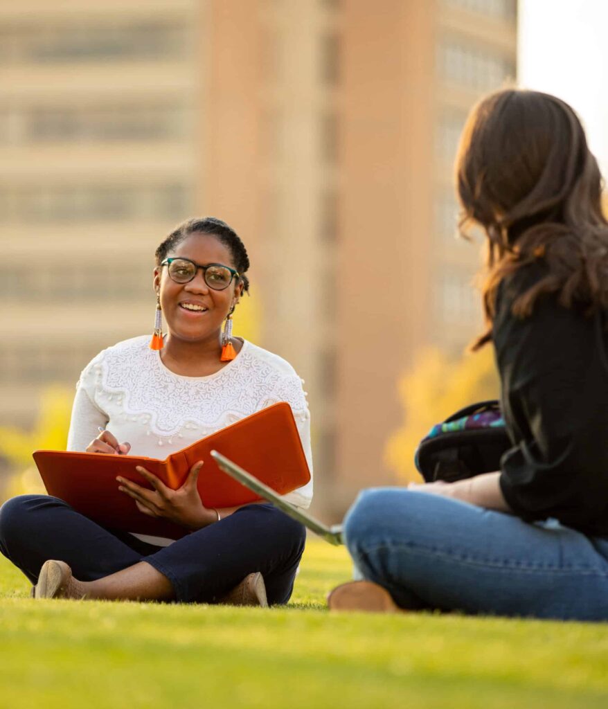 A student sits cross-legged on the Humanities lawn with an orange folder open in her lap, talking with another student