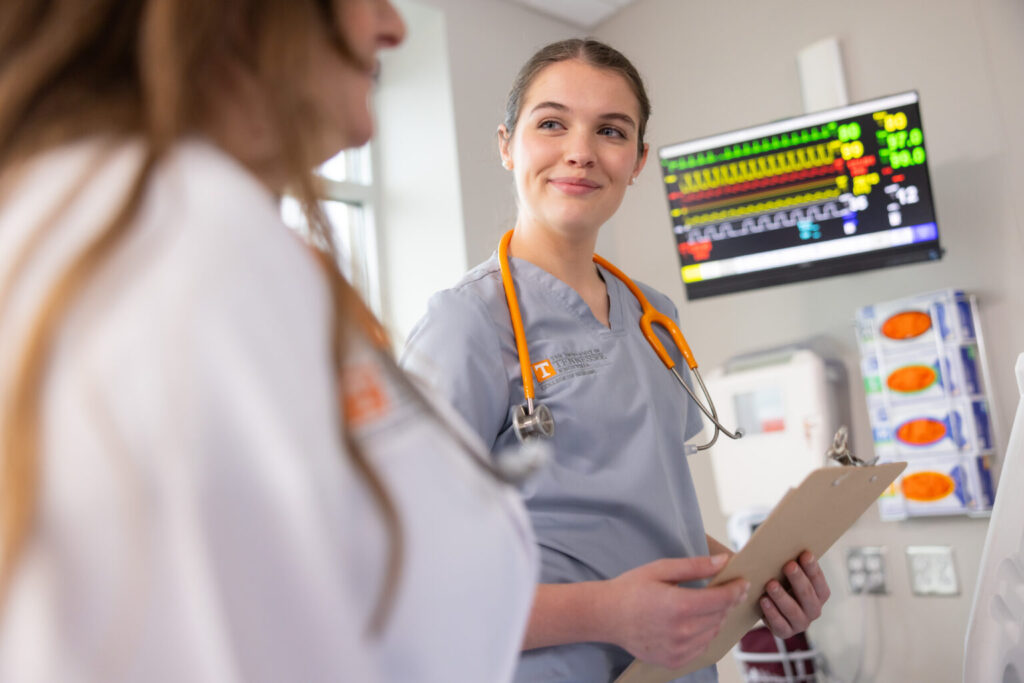 A nursing student with an orange stethoscope around her neck, standing in a room with diagnostic panels on the wall