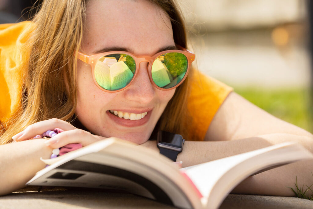 A UT student laying outside reading a book in the sunlight, wearing orange sunglasses