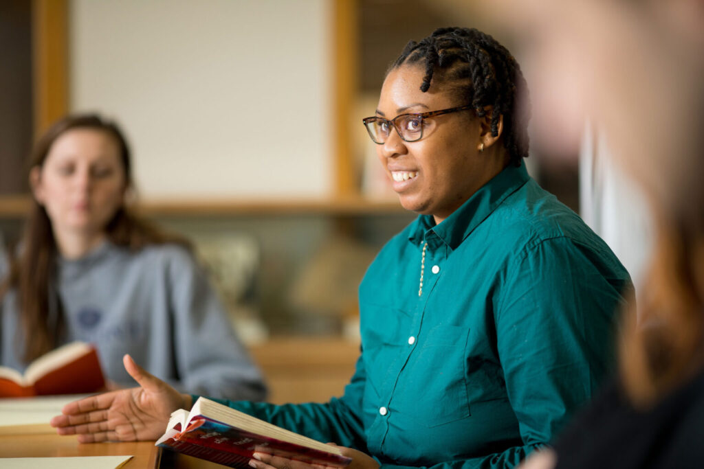 A classroom shot of a person wearing a teal shirt and talking with classmates while holding a book