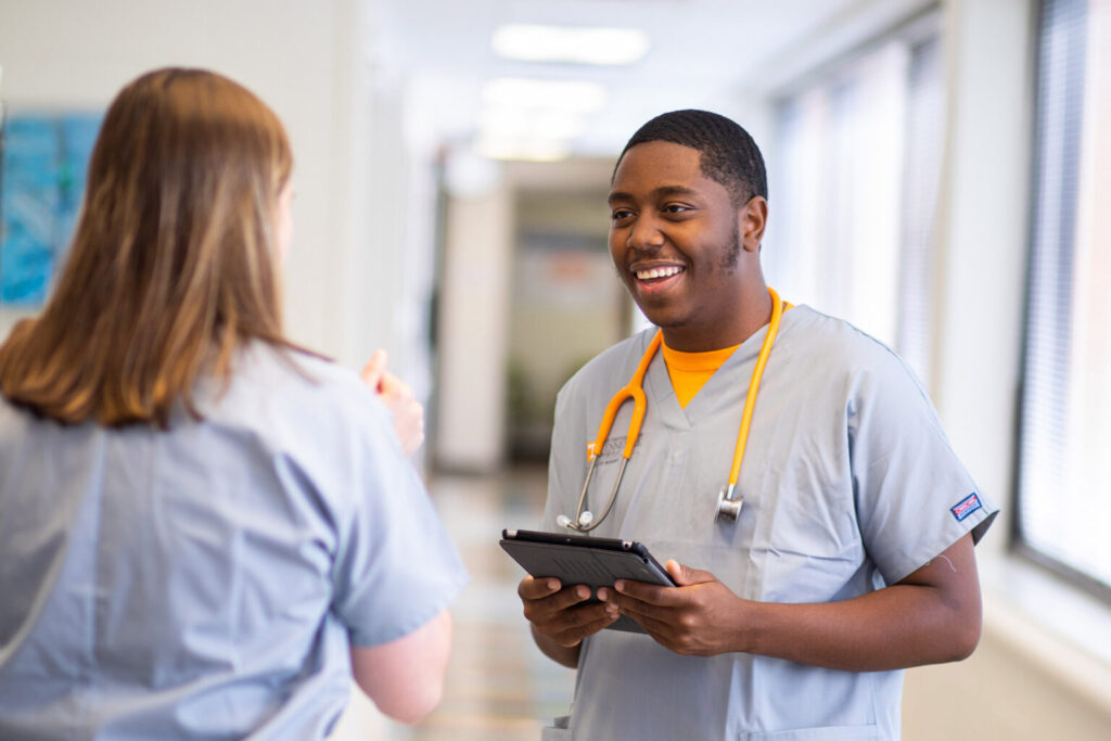A smiling nursing student, wearing scrubs and an orange stethoscope draped around his neck, speaks with another nurse in a hallway. Her back is to the camera.