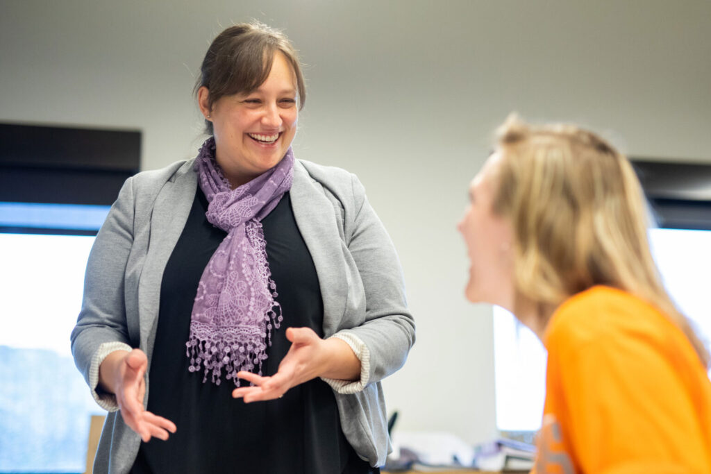 A professor wearing a gray cardigan and purple scarf gestures as she talks with a seated student, who is wearing an orange shirt.