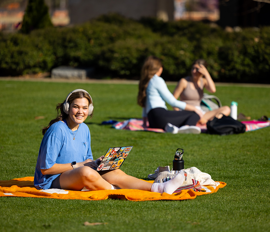 A student smiles for the camera while working on a laptop and laying on a UT blanket at the Humanities Plaza-North lawn