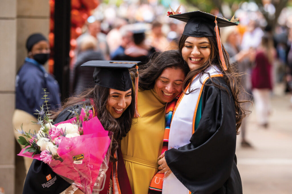 Students wearing caps and gowns and holding bouquets of flowers embrace following the commencement ceremony