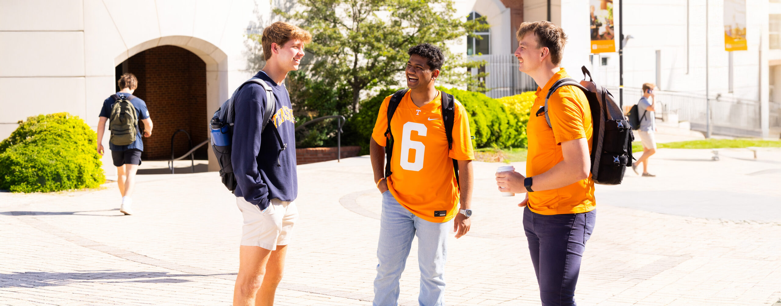 Three UT students wearing Tennessee gear and backpacks stand outside in the sunlight smiling and chatting