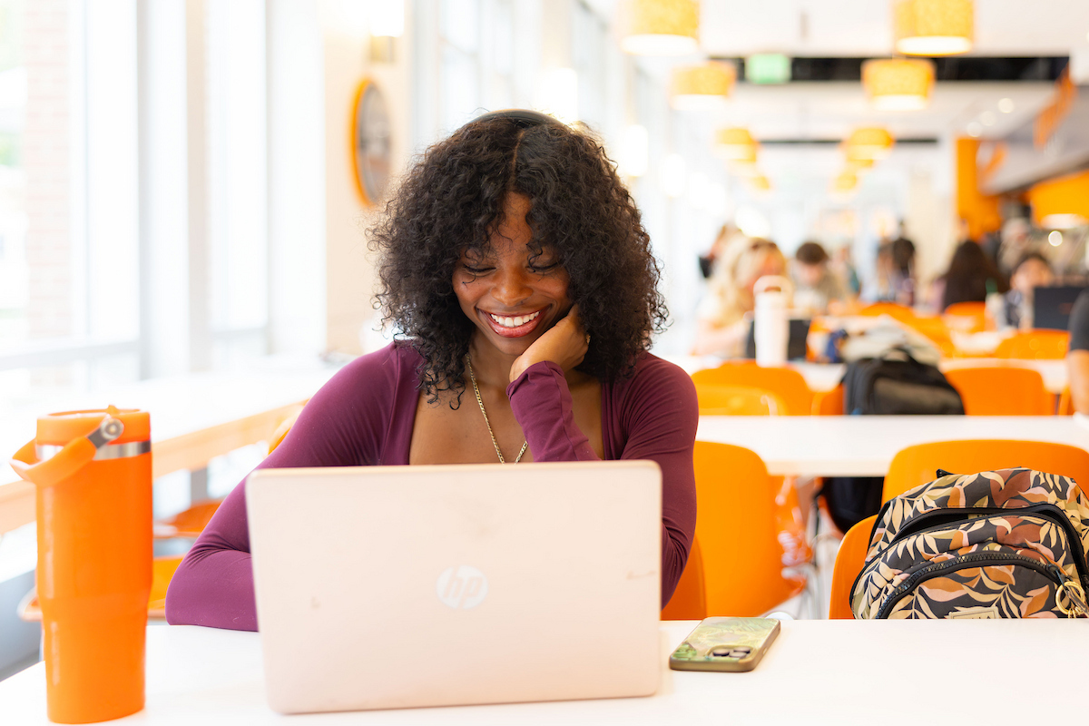 A person with curly hair smiles while using a laptop at a table in a brightly lit, modern cafeteria.
